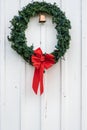 Christmas wreath made of artificial evergreen boughs and a light string, with red bow and cow bell, on a white wall Royalty Free Stock Photo
