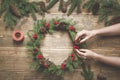 Christmas wreath made of spruce branches with holly berries on wooden board. Female hand decorated wreath. Top view. Royalty Free Stock Photo