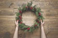 Christmas wreath of spruce branches with holly berries on wooden board. Female hand holding wreath. Top view. Royalty Free Stock Photo