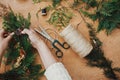Christmas wreath workshop. Hands holding fir branches, pine cones, berries, thread, scissors on wooden table, flat lay. Making Royalty Free Stock Photo