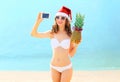 Christmas young smiling woman in red santa hat with pineapple taking picture self portrait on smartphone at beach over sea