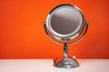 Chromed steel round magnifying mirror on handle set up on the white table. Bright orange background, single object.