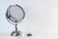 Chromed steel round magnifying mirror on handle set up on the white table with some jars with creams and cosmetics. Cold light blu