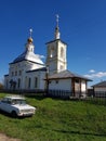 The church is white in color behind an iron fence. A white car near the church. Green grass. Two crosses of gold in a blue sky.