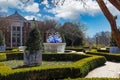 A circular stone water fountain with curve blue and white glass sculptures surrounded by lush green trees with a gorgeous blue sky