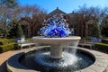 A circular stone water fountain with curve blue and white glass sculptures surrounded by lush green trees with a gorgeous blue sky