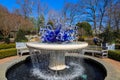 A circular stone water fountain with curve blue and white glass sculptures surrounded by lush green trees with a gorgeous blue sky