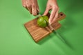 A woman`s hands cut a natural green ripe lime in half on a wooden cutting board on a green background with copy space. Royalty Free Stock Photo