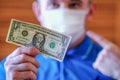 A dollar in the hand of a man with a protective mask on his face. The concept of disparity between the price of life and the price