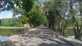 In the city park, the asphalted road across the river passes over a concrete bridge with metal railings. Concrete tile sidewalks a