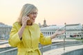 City portrait of mature smiling woman in glasses, yellow coat talking on mobile phone, background urban panorama, copy space