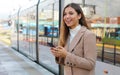 City transport. Happy beautiful woman holding cellular at tram stop. Smiling business woman satisfied with online ticket service