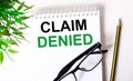 CLAIM DENIED is written in a white notebook next to a pencil, black-framed glasses and a green plant Royalty Free Stock Photo