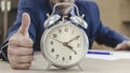 Classic alarm clock on the office table. Against a blurred background, the boss shows a thumb gesture. Completed task on time.