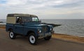 Classic Blue Land Rover being driven along seafront promenade ocean in background.