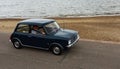 Classic Blue Leyland Mini being driven along seafront promenade ocean in background.