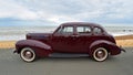 Classic Dark Red Studebaker motor car parked on seafront promenade beach and sea in background.