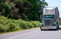 Classic green big rig semi truck with chrome pipes and bulk trailer for commercial cargo driving by green road with trees Royalty Free Stock Photo