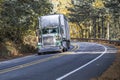 Classic green big rig semi truck tractor with day cab transporting covered bulk semi trailer driving on the narrow winding road in Royalty Free Stock Photo