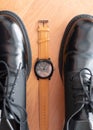 Classic mechanical wrist watch lay between pair of man black formal shoes. top view