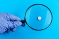 Classic medical pill with risk, doctor`s hand in gloves and magnifying glass in blurred foreground. Blue background