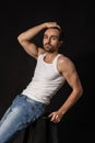 Classic portrait of young handsome bearded sad man in white undershirt and jeans isolated on dark background. Emotions Royalty Free Stock Photo