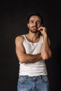 Classic portrait of young handsome bearded sad man in white undershirt and jeans isolated on dark background. Emotions Royalty Free Stock Photo
