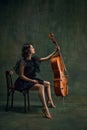 Classical music live performance. Elegant young woman, cellist, passionate musician sitting with cello against dark Royalty Free Stock Photo