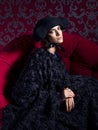 Classical portrait of woman wearing black hat end dress sitting on red sofa