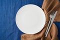 Clean empty white plate, fork and knife on bright blue dark linen tablecloth on table, copy space Royalty Free Stock Photo