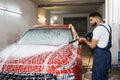 Handsome young bearded man worker, wearing protective clothes and gloves, washing modern red car Royalty Free Stock Photo