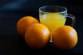 A clear glass mug filled with orange juice and three fresh oranges stand on a dark surface