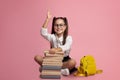 Clever pupil. Smiling girl in glasses with stack of books and backpack knows answer and raises hand up Royalty Free Stock Photo