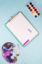 Clipboard with blank paper sheets,multicolored watercolor paints in box, paint brushes and palette on blue table. Top Royalty Free Stock Photo