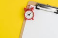 Clipboard with empty white paper with read alarm clock on solid yellow background with copy space for writing message using as
