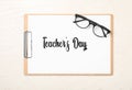 Clipboard with words TEACHER`S DAY and glasses on white wooden background, top view