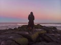 cloaked figure staring out over the ocean from a jetty at sunset