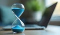 Close photo of blue sand hourglass. Sand flows down representing passing time. Blurred laptop on table in the background. Concept Royalty Free Stock Photo