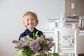 Close portrait of a beautiful blond child, toddler boy holding vase with lilacs at home, putting it on the table to decorate