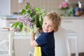 Close portrait of a beautiful blond child, toddler boy holding vase with lilacs at home, putting it on the table to decorate