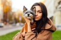 Close portrait of a woman with a biewer terrier dog in her arms on the street. The owner hugs a small dog on a walk and looks at