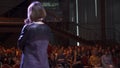 Close-up of adult female speaker with short hair in a grey dress standing on stage and reporting for audience. Art