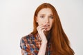 Close-up of anxious young woman with ginger hair, biting finger and staring nervous at camera, having worries, standing Royalty Free Stock Photo