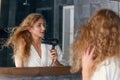 Close up of appealing smiling young lady in white dressing gown posing on camera while drying her curly hair using hair Royalty Free Stock Photo