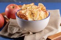 Close-up apple chips in cup and fresh apples on beige fabric on wooden tray on blue background