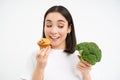 Close up of asian female model, eating cupcake, bite pastry and looking at healthy vegetable broccoli, white background Royalty Free Stock Photo