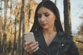 Close up of attractive brunette girl holding smart phone at unfocused forest background. Female can`t stop checking her