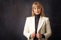 Close-up of an attractive woman smiling and looking at camera. Blond haired female wearing blazer and standing against dark