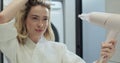 Close up of attractive young woman looking at her reflection in the mirror and drying hair with blow dryer in bathroom Royalty Free Stock Photo