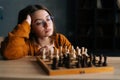 Close-up of attractive young woman wearing elegant eyeglasses thinking about chess move while sitting on wooden floor in Royalty Free Stock Photo